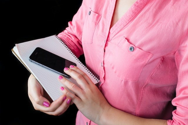 Woman in pink button down shirt holding phone and notebook